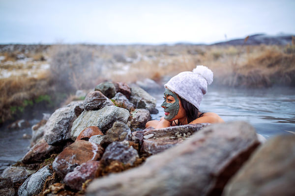 woman soaking in hot spring with mud mask and puff wool hat on